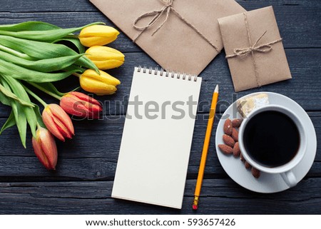 Notepad with pencil next to the tulips, coffee, and envelopes on wooden background. View from above with copy space.