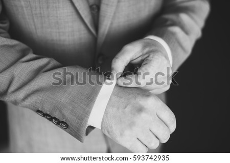 Elegant young fashion man dressing up for wedding celebration. Color close up image of male hands. Groom buttoning a shirt. Dark background. Black and white photo