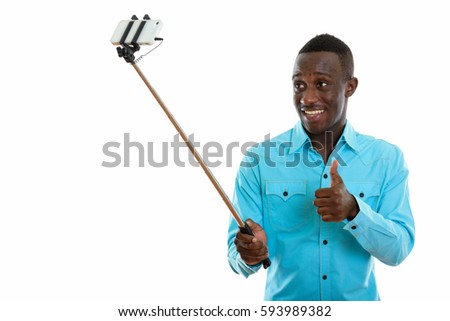 Studio shot of young happy black African man smiling and giving thumb up while taking selfie picture with mobile phone on selfie stick