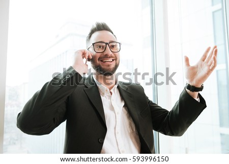 Happy Bearded business man in suit and eyeglasses which talking on phone with window on background