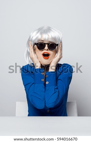 blonde in blue shirt, red lips in black glasses smiling on a light background
