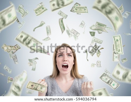 Portrait of a stressed out blond woman yelling and clenching her fists near a gray wall while standing under a dollar rain. Concept of bankruptcy