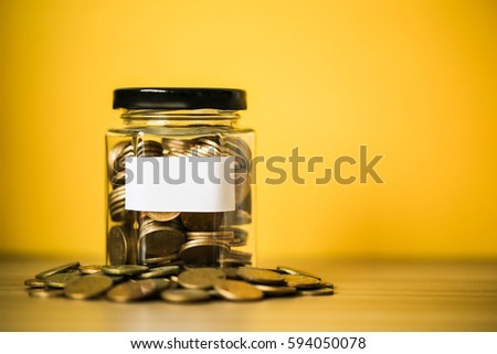 A lot coins in glass money jar with blank white paper on yellow background.