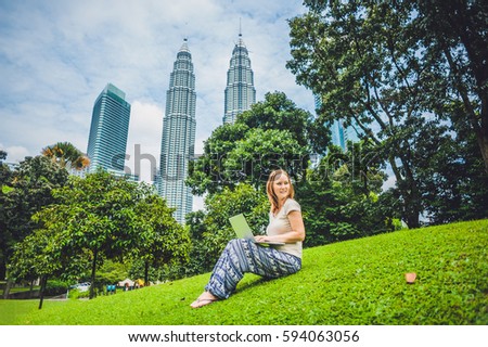 A young woman in casual dress using laptop in a tropical park on the background of skyscrapers. Mobile Office concept.