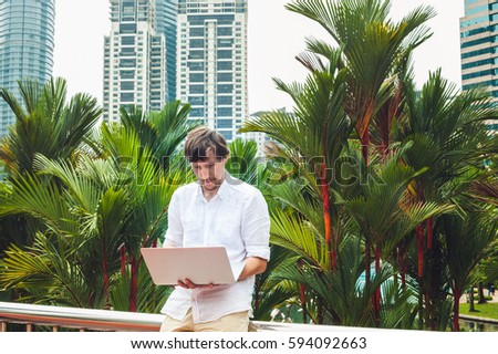 Man businessman or student in casual dress using laptop in a tropical park on the background of skyscrapers. Dressing in a white shirt, beige shorts. Mobile Office concept.