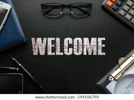 Black chalkboard with business accessories (notepad, magnifying glass, fountain pen, tablet, phone, glasses and calculator) and text WELCOME. Top view.