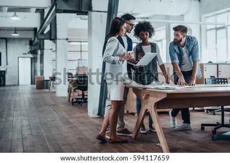 Working through some concepts. Group of young business people working together in creative office while standing near the wooden desk   Royalty-Free Stock Photo #594117659