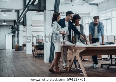 Passionate about their project. Group of young business people working together in creative office while standing near the wooden desk   Royalty-Free Stock Photo #594117701