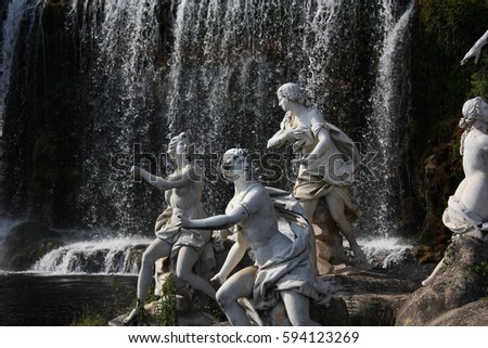 Group of old statues of goddesses in an artificial lake with a waterfall in the background.