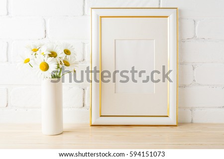 Gold decorated frame mockup with wild daisy flower in styled vase near painted brick wall. Empty frame mock up for presentation design.  Template framing for modern art. 