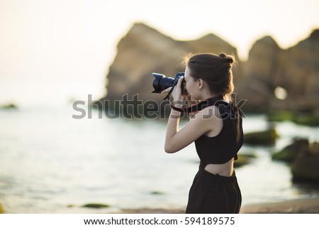 Young woman photographer holding camera and looking into frame. Girl dressed in black jumpsuit, European appearance. Concept of feminine beauty,  work photographer tourist with camera on vacation o