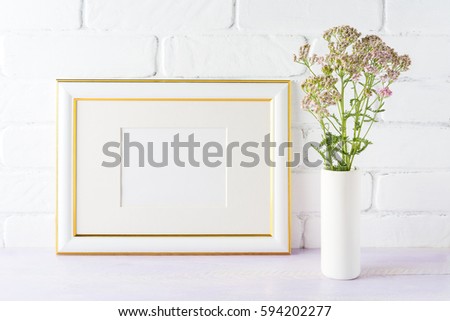 Gold decorated landscape frame mockup with wild creamy pink flowers in cylinder vase near painted brick wall. Empty frame mock up for presentation design.  Template framing for modern art.  