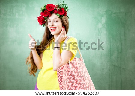 Colorful portrait of a beautiful pregnant woman in yellow t-shirt with wreath made of red roses holding shopping bag on the green background
