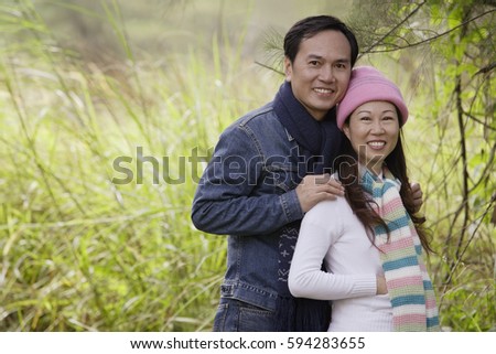 Man and woman wearing hat and scarves, standing outdoors in nature, smiling at camera