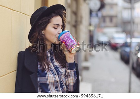 Outdoor portrait of young attractive lady with curly brunette hair in early spring. Model wearing a coat, checkered shirt and a hat and drinking coffee to go.