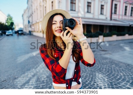 Lovely tourist girl with brown hair wearing hat and red shirt, making photo with camera at old European city background and smiling, traveling.