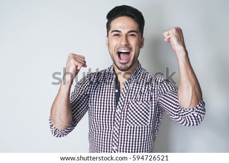 Yes I'm winner! Pleased and happy young brunette man in checkered shirt is smiling broadly and showing victory sign with both hands against white background