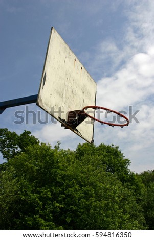 Basketball hoop with backboard but missing the net. Trees and blue sky with white cloud in the background.