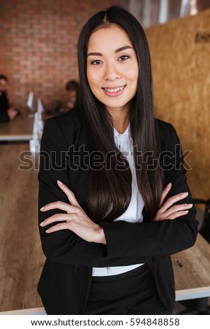 Smiling attractive asian young businesswoman standing with hands folded in office