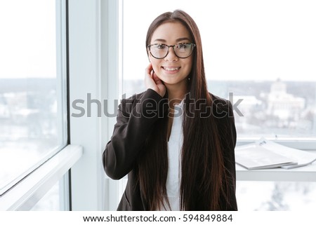 Cheerful asian young businesswoman in glasses standing near the window in office