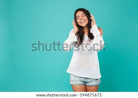 Cheerful pretty young woman in headphones standing and listening to music over blue background