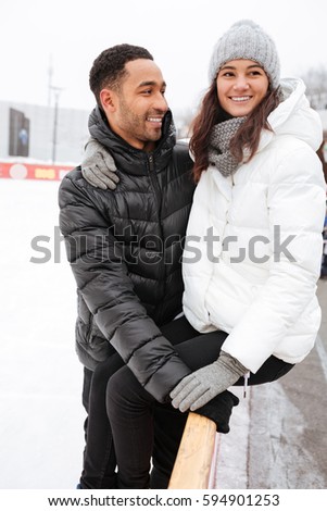 Photo of young attractive loving couple hugging and skating at ice rink outdoors. Looking aside.