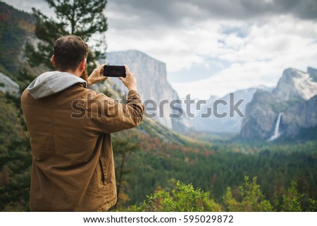 man in brown coat taking photo of yosemite with smartphone