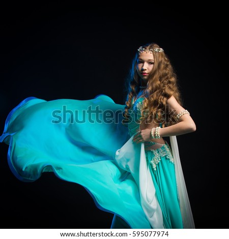 Portrait of a young girl with long hair in a turquoise costume oriental dancer dancing on a black background in the scenic blue light