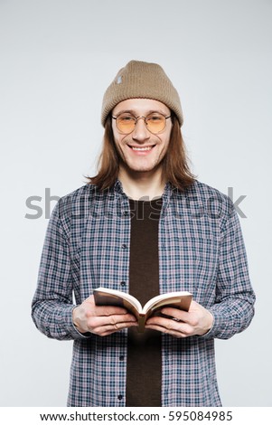 Vertical image of hipster in eyeglasses which posing in studio with book and looking at camera. Isolated gray background