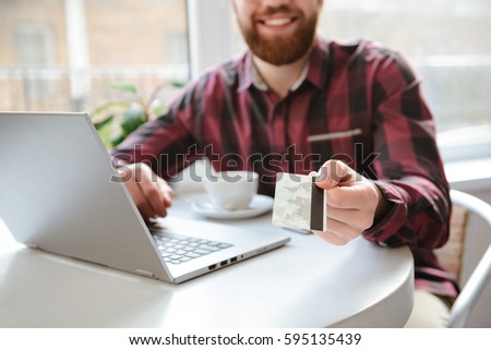 Cropped photo of cheerful bearded young man sitting in cafe while using laptop computer and showing debit card to camera.