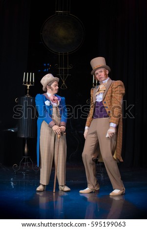 Two male actors in old frocks and hats cylinders posing against the backdrop of scenery for the performance