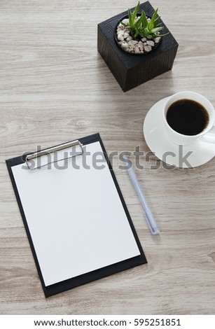 Workplace. On the wooden table is a folder with a clip, a white sheet with space for text, pot with plant, a pan and white cup of coffee.