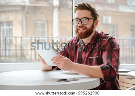 Picture of happy bearded young man student wearing glasses sitting in cafe while using tablet computer. Looking aside.