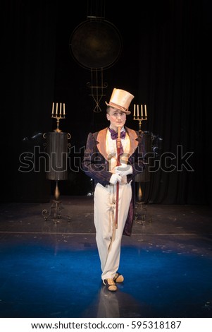 A male comedian in an old frock coat and a hat of a cylinder poses against the backdrop of the scenery for the play