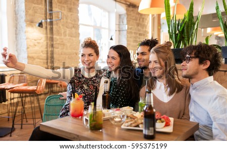 leisure, technology, friendship, people and holidays concept - happy friends with food and drinks taking selfie by smartphone at bar or cafe