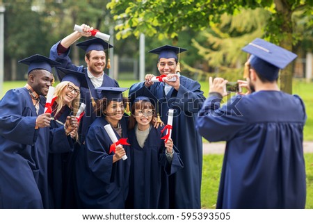 education, graduation, technology and people concept - group of happy international students in mortar boards and bachelor gowns with diplomas taking picture by smartphone outdoors