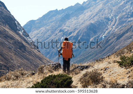 Rear View of Hiker with large orange Backpack covered by waterproof cover staying on grassy trail and overlooking Mountain Valley