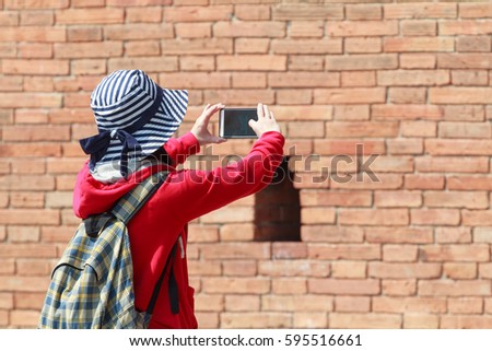 Woman Tourist is Taking Photo by Mobile Phone with Big Brick City Wall Background. Set as Copy Space for Text.