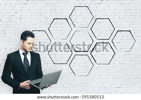 Handsome young businessman using laptop on brick wall background with abstract drawn cells. Copy space. Technology concept
