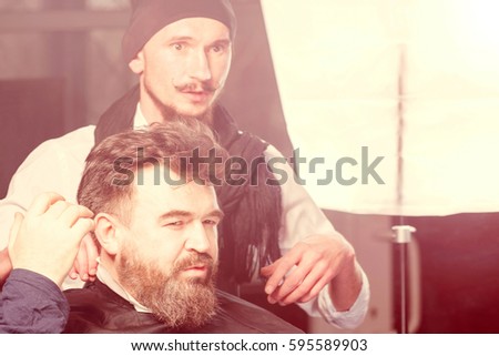 Barber in a hat and a black apron with a comb in his hand and sitting in front of him brunette man with a beard