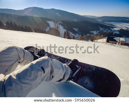 Snowboard and boots of a man sitting on a skiing trail with view down the hill on a track and distant mountains in a sunny winter day