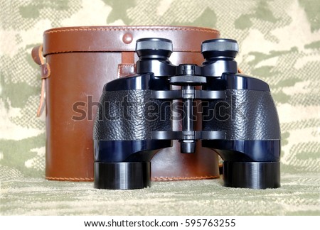 Vintage military Porro prism black color binoculars with brown leather carry case with strap on camouflage background front view close up