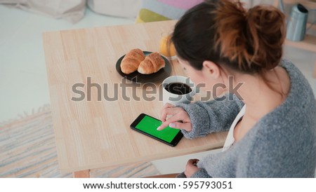 Young woman sitting at the table and drinking coffee in kitchen. Brunette girl uses smartphone, green screen.