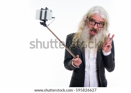 Studio shot of happy senior bearded businessman smiling and giving peace sign while taking selfie picture with mobile phone on selfie stick