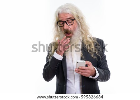 Studio shot of senior bearded businessman looking shocked while using mobile phone