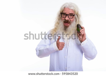 Studio shot of happy senior bearded man doctor smiling while giving thumb up and holding avocado