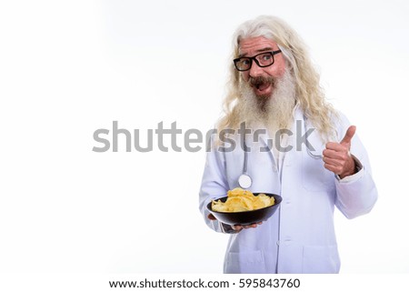 Studio shot of happy senior bearded man doctor smiling while giving thumb up and holding bowl of potato chips
