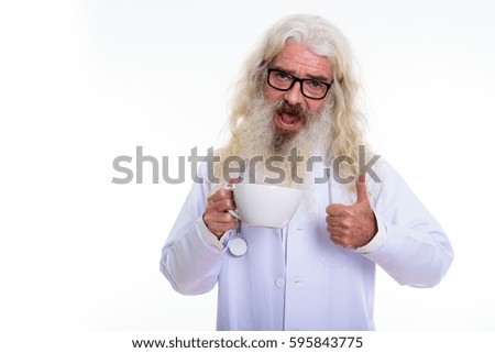 Studio shot of happy senior bearded man doctor smiling while giving thumb up and holding coffee cup