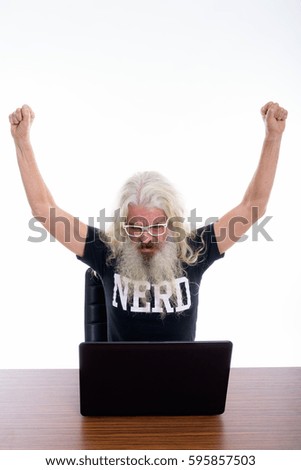 Studio shot of senior bearded nerd man raising both arms and looking excited while using laptop on wooden table
