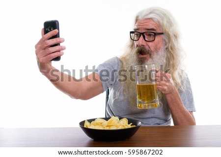 Happy senior bearded man smiling while taking selfie picture with mobile phone while holding glass of beer with bowl of potato chips on wooden table